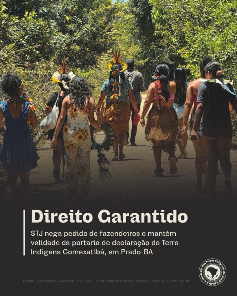 Card informativo com uma fotografia de um grupo de pessoas indígenas caminhando por uma estrada cercada por vegetação densa. Algumas pessoas usam cocares de penas, saias de fibras vegetais, adornos tradicionais e carregam objetos cerimoniais, enquanto outras vestem roupas casuais. A imagem é vista por trás do grupo, que segue em direção ao interior da mata. Na parte inferior, sobre um fundo escuro, aparece o título em destaque "Direito Garantido". Abaixo, lê-se: "STJ nega pedido de fazendeiros e mantém validade da portaria de declaração da Terra Indígena Comexatibá, em Prado-BA". No canto inferior direito está o logotipo da Articulação dos Povos Indígenas do Brasil (APIB), e na base do card aparecem os nomes de organizações indígenas apoiadoras.