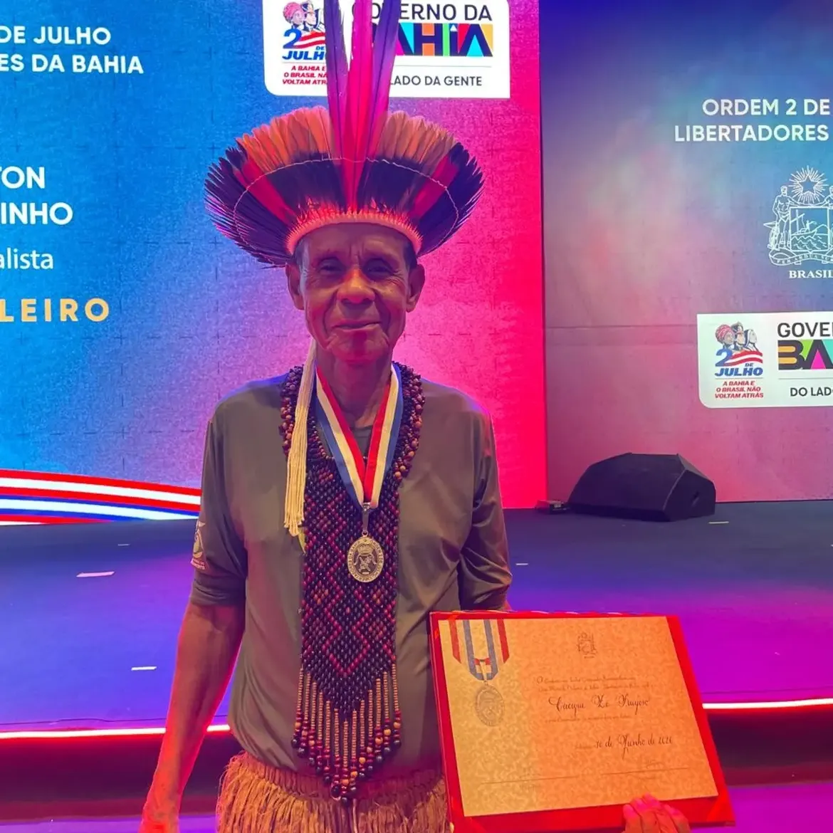 Fotografia do cacique Zé Fragoso em cerimônia de condecoração da Ordem 2 de Julho – Libertadores da Bahia. Ele aparece em pé, sorrindo, usando cocar de penas vermelhas, azuis e pretas, colares tradicionais, vestimenta indígena e a medalha da honraria ao redor do pescoço. Em uma das mãos, segura o diploma da condecoração. Ao fundo, há um palco iluminado com as cores azul e vermelha, além de painéis com a identidade visual do Governo da Bahia e da Ordem 2 de Julho.