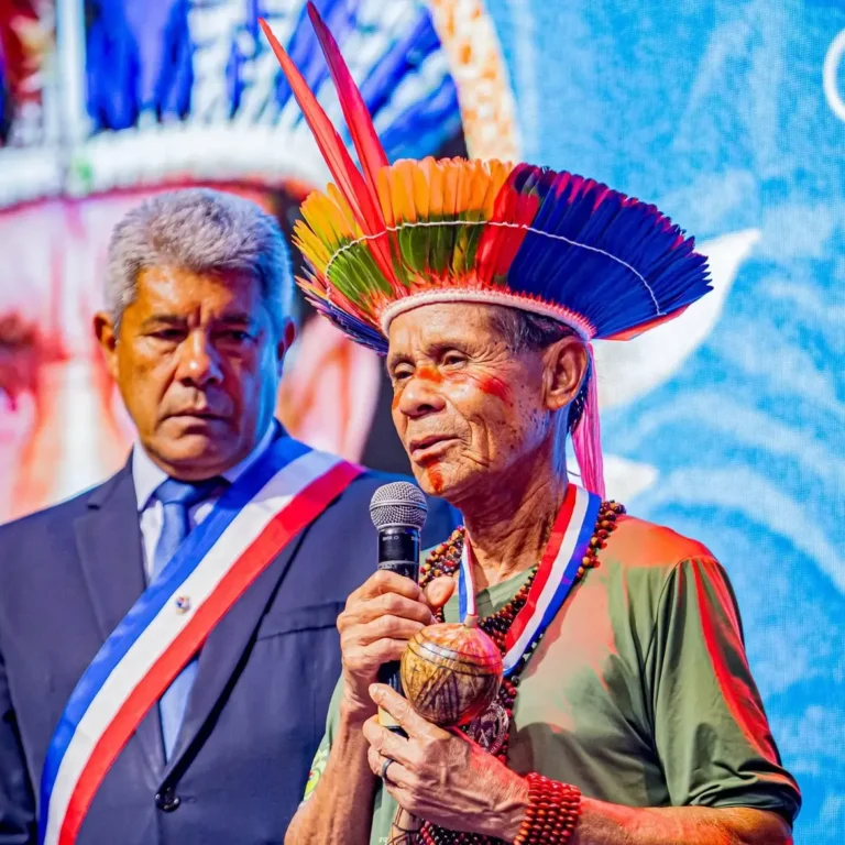 O cacique Zé Fragoso discursa ao microfone durante a cerimônia de concessão da Ordem 2 de Julho – Libertadores da Bahia. Ele usa um cocar colorido de penas, pinturas faciais vermelhas, colares de sementes e uma medalha no pescoço, enquanto segura um maracá nas mãos. Ao fundo, o governador da Bahia, Jerônimo Rodrigues, vestido de terno e usando uma faixa cerimonial, acompanha o discurso em um palco iluminado por um grande painel de LED.