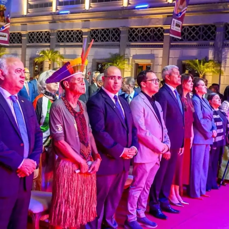 O cacique Zé Fragoso está em pé na primeira fila da cerimônia de concessão da Ordem 2 de Julho – Libertadores da Bahia, usando cocar de penas coloridas, colares de sementes, camiseta verde e saia de fibras naturais. Ao seu lado, o governador da Bahia, Jerônimo Rodrigues, e outras autoridades acompanham o evento de pé. A cerimônia acontece em um espaço histórico iluminado com luzes coloridas, diante de um palco.