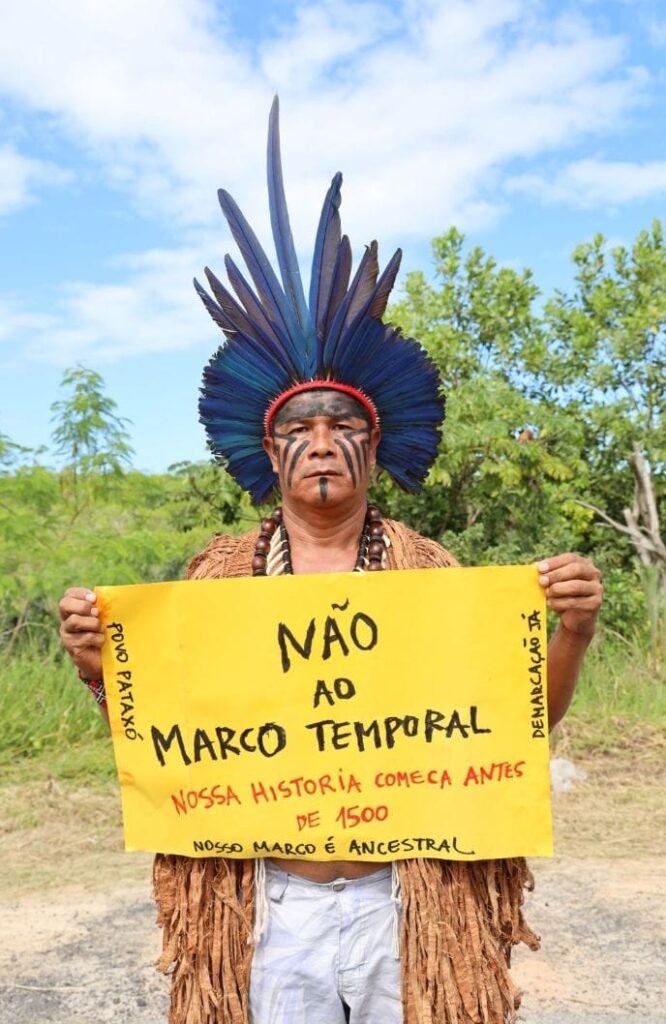 O cacique Louro Pataxó está em pé ao ar livre, diante de uma área de vegetação e céu azul com nuvens, usando um grande cocar de penas azuis, colares e vestimenta tradicional. Seu rosto está pintado com grafismos pretos. Ele segura um cartaz amarelo com a mensagem: “Não ao Marco Temporal. Nossa história começa antes de 1500. Nosso marco é ancestral”. Nas laterais do cartaz aparecem as inscrições “Povo Pataxó” e “Demarcação Já”. A imagem expressa mobilização em defesa dos direitos territoriais indígenas e da demarcação de terras.