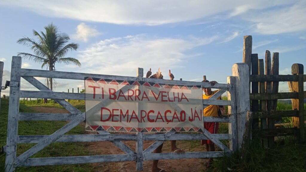 Fotografia de uma porteira de madeira branca em uma área rural, com uma faixa presa ao centro onde se lê: "T.I. Barra Velha – Demarcação Já!" em letras vermelhas. Atrás da porteira, algumas pessoas aparecem parcialmente visíveis, fazendo sinal de positivo com as mãos. Ao fundo há um coqueiro, vegetação, cercas de madeira, um caminho de terra e um céu azul com nuvens iluminadas pela luz do fim da tarde.