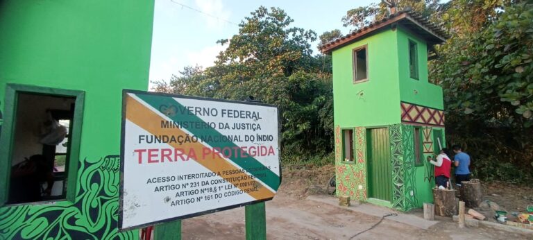 Fotografia da entrada da Aldeia Velha, em área cercada por vegetação. Em primeiro plano, há uma placa com os dizeres “Governo Federal”, “Ministério da Justiça”, “Fundação Nacional do Índio” e “Terra Protegida”, informando a restrição de acesso a pessoas estranhas à comunidade. Ao fundo, observam-se construções pintadas em verde, decoradas com grafismos indígenas em preto, vermelho e branco. À direita, duas pessoas realizam trabalhos de pintura na fachada de uma guarita de dois pavimentos.