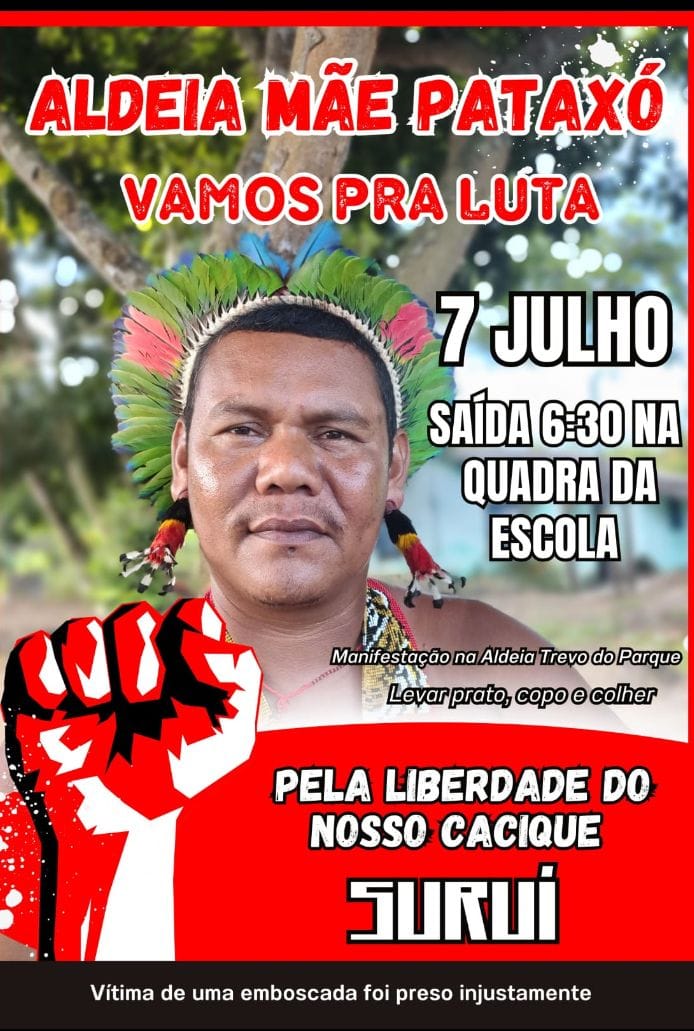 Cartaz de mobilização da comunidade indígena Pataxó. No centro, há a fotografia de um homem indígena, o cacique Suruí usando cocar colorido, com penas verdes e vermelhas. No topo, em letras grandes e vermelhas, lê-se: "Aldeia Mãe Pataxó – Vamos pra luta". À direita da foto, em letras pretas: "7 julho – Saída 6:30 na quadra da escola". Abaixo, há um punho fechado estilizado em vermelho, preto e branco, símbolo de resistência. Em destaque, aparece o texto: "Pela liberdade do nosso cacique Suruí". Na parte inferior, em faixa preta, está escrito: "Vítima de uma emboscada foi preso injustamente". Também consta a instrução: "Manifestação na Aldeia Trevo do Parque – Levar prato, copo e colher"